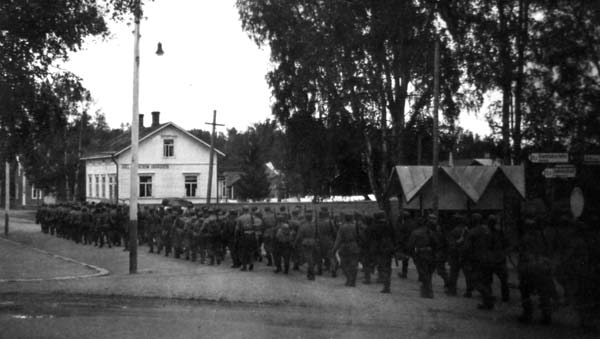 Kanske någon av de som skickade hälsningar finns bland dessa soldater som 1941 marscherade iväg mot Kovjoki för vidare transport till fronten. Från korsningen esplanaden–Bankgatan mot ostsydost. På byggnaden i fonden, nuvarande Folkhälsan, står ”JOEL FINNSTRÖM SKRÄDDERI”.