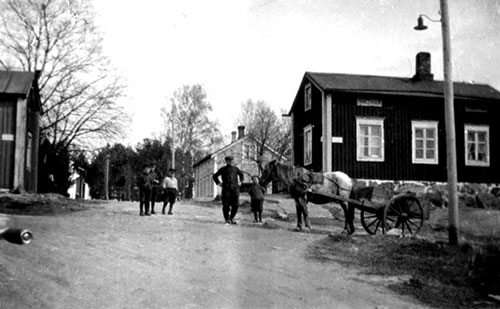 Korsningen Karleborgsgatan - Källbackgatan från väster. Andells gård i bakgrunden. Notera cykelhandtaget i vänsterkant! F.d. Widmans till vänster och f.d. Melchers till höger. Förstoring.