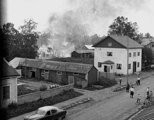 Mattssons/Spåras uthus brinner somamren 1957. Mannen som står med keps och spanar är Rolf Jansson. Blomströms uthus mitt i bild , KAP-PAS till höger och Bönehuset i högerkant. Förstoring.