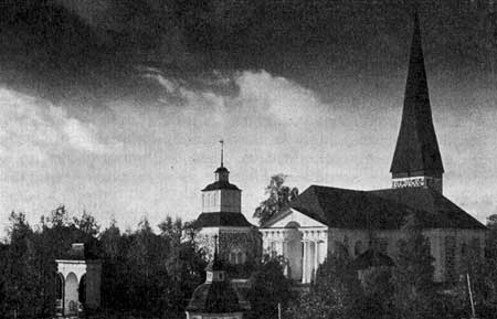 Pedersöre kyrka och klockstapel med stigport och benhus i förgrunden.